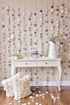 a white table topped with a cake next to a wall covered in heart shaped paper garlands