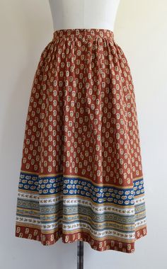 "This gorgeous skirt is gently gathered at the waist and comes in a handsome shade of rust--somewhere at the nexus of brown, red, and orange. The print features golden yellow paisley-like motifs as well as ornate bands of white and navy blue with pale green accents. Zippered back closure (nylon coil teeth) with an attractive plastic button. Good, wearable vintage condition. One tiny hole on the front left about the size of a pinhead (pictured), which appears to be a burn mark. We spotted two or Bohemian Brown Pleated Maxi Skirt, Brown Vintage Tiered Skirt, Vintage Brown Tiered Skirt, Vintage Brown Flowy Skirt, Vintage Cotton Brown Skirt, Traditional Long Brown Skirt, Vintage Skirt With Elastic Waistband In Relaxed Fit, Vintage Relaxed Skirt With Elastic Waistband, Vintage Long Skirt With Elastic Waistband