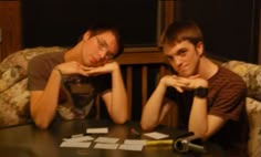 two young men sitting at a table in front of each other with their hands on their chins