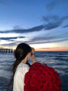 a woman holding a bouquet of red roses in front of the ocean with a pier in the background