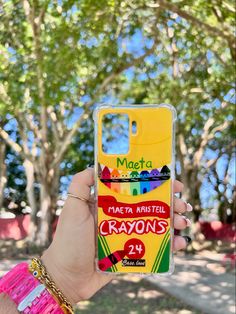 a person holding up a phone case with crayons on it in front of trees