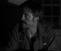 a man with suspenders and a mustache stares into the distance in a dark room