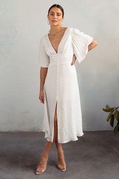 a woman standing in front of a wall wearing a white dress with ruffle sleeves