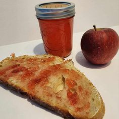 a piece of bread next to an apple and a jar of jam on a table