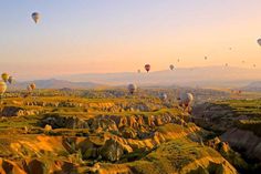 many hot air balloons flying in the sky over some hills and valleys with mountains in the background