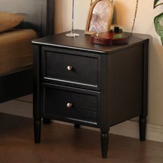 a black night stand with two drawers in front of a bed and a mirror on the nightstand