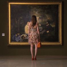 a woman is standing in front of a painting and holding an orange purse while looking at it