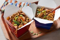 two takeout boxes filled with noodles and meat on a wooden platter next to chopsticks