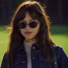 a woman wearing sunglasses and a denim jacket looks off to the side while standing in front of a grassy area