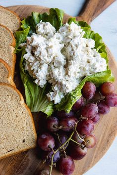 a wooden plate with bread, grapes and chicken salad on it next to some slices of bread