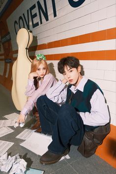 two young people sitting on the ground in front of a wall with paper all over it