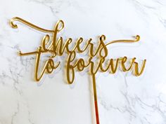 there is a cake topper that says cheers to forever in gold letters on a marble surface