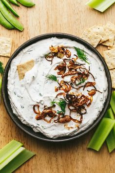 a bowl filled with white dip surrounded by celery and crackers on a wooden table