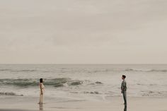 a man and woman standing on the beach next to the ocean looking at each other