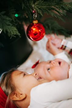 two babies laying under a christmas tree with ornaments hanging from it's branches and one is looking at the camera