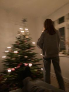 a woman standing next to a christmas tree in a living room with lights on it