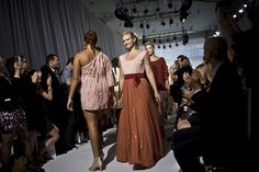 two women walking down the runway at a fashion show with people taking pictures behind them