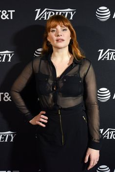 a woman with red hair standing in front of a black wall and wearing a sheer shirt