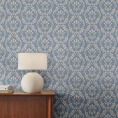 a lamp on a table next to a blue wallpaper with white floral designs and a wooden cabinet