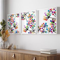 three paintings on the wall above a dresser with flowers in vases next to it