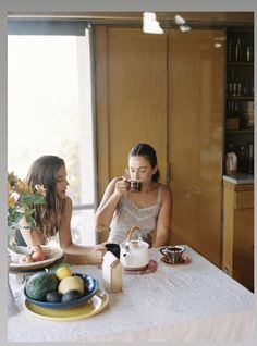 two women sitting at a table with tea and fruit in front of them, talking