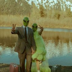 two people dressed in alien costumes standing next to a body of water with suitcases