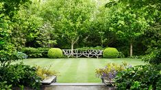 a lush green yard surrounded by trees and bushes with benches on each side, in front of a wooden bridge