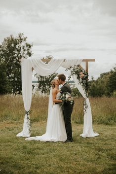 a bride and groom kissing under an arch decorated with greenery at their outdoor wedding ceremony