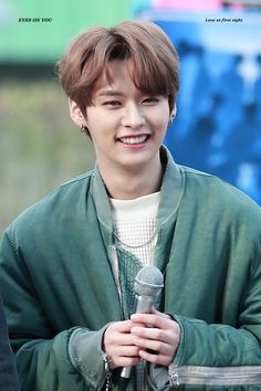 the young man is holding a microphone in his hand and smiling at the camera while wearing a green jacket