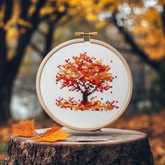 a cross - stitch tree is displayed on a wooden stump in the fall with leaves around it