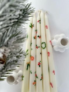 four white candles with holly designs on them next to a christmas ornament and pine tree