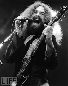 a man with long hair and beard holding a guitar in front of a microphone on stage