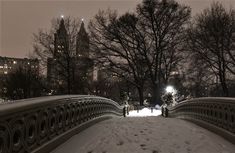 a bridge that has snow on the ground and trees in the background with buildings lit up at night