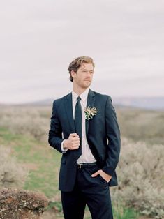 a man wearing a suit and tie standing in front of a rock formation with his hands on his hips