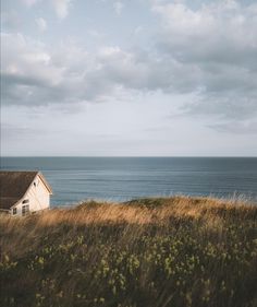 a house sitting on top of a grass covered hillside next to the ocean with clouds in the sky