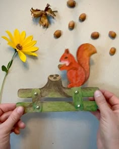 two hands are holding a piece of wood with a squirrel on it and flowers in the background