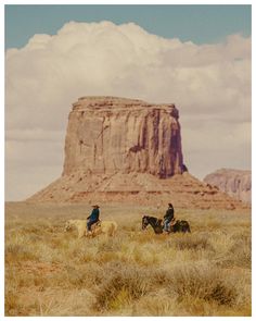 two people riding horses in the desert with a large rock formation in the back ground