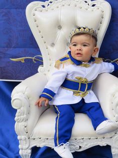 a baby sitting in a white chair wearing a blue and gold outfit with a crown on it's head