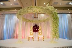 a stage set up for a wedding ceremony with white flowers and gold chairs on it