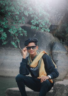 a young man sitting on top of a rock next to a river wearing sunglasses and a scarf