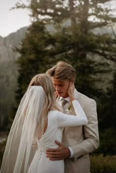 a bride and groom embracing each other in front of some trees with mountains behind them