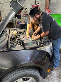 two people working on a car engine in a garage with the hood open and one person standing next to it