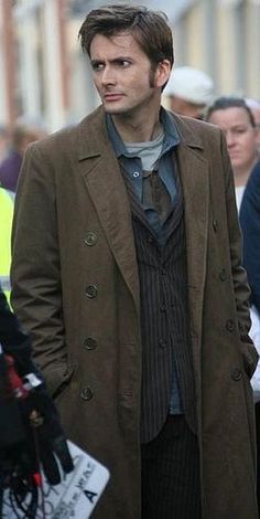 a man in a trench coat and tie standing on the street with other people behind him