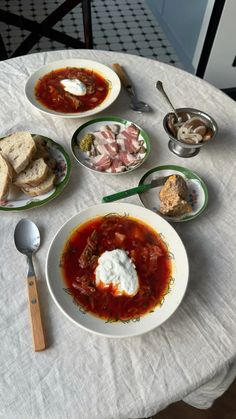 a table topped with plates of food next to bowls of soup and bread on top of a white table cloth