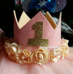 a pink princess crown with gold glitter number one on it's side and flowers around the edges