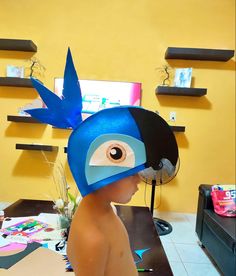 a child wearing a blue bird mask with an eye on it's head, in front of a yellow wall