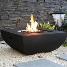 a fire pit sitting on top of a gravel field next to a stone wall and water fountain