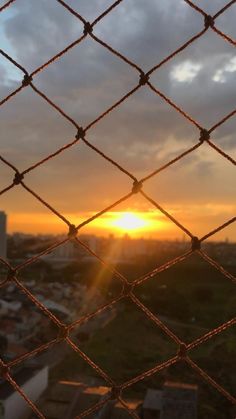 the sun is setting behind a chain link fence with buildings in the backgroud