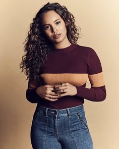 a woman with curly hair wearing jeans and a sweater is posing for the camera while holding her hands in her pockets