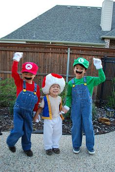 three children dressed up as mario and luigi in front of a fence with their arms in the air